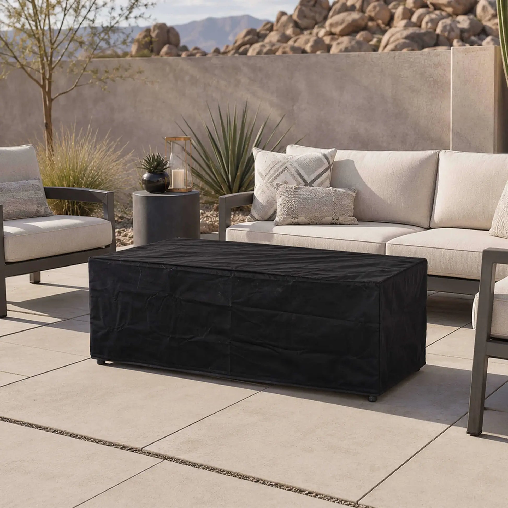 Flame Havens Refuge Series 68 x 30 x 24-inch rectangular gray propane fire pit protected by a fitted black canvas fabric cover on a modern desert patio, surrounded by cushioned outdoor seating, decorative plants, boulders, and mountain views.