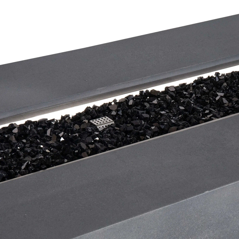 Detailed close-up of the Flame Havens Refuge Series 68 x 30 x 24-inch gray GFRC concrete propane fire pit, showing glossy black reflective fire glass spread across the linear stainless steel burner pan with the perforated ignition cage visible near the center.