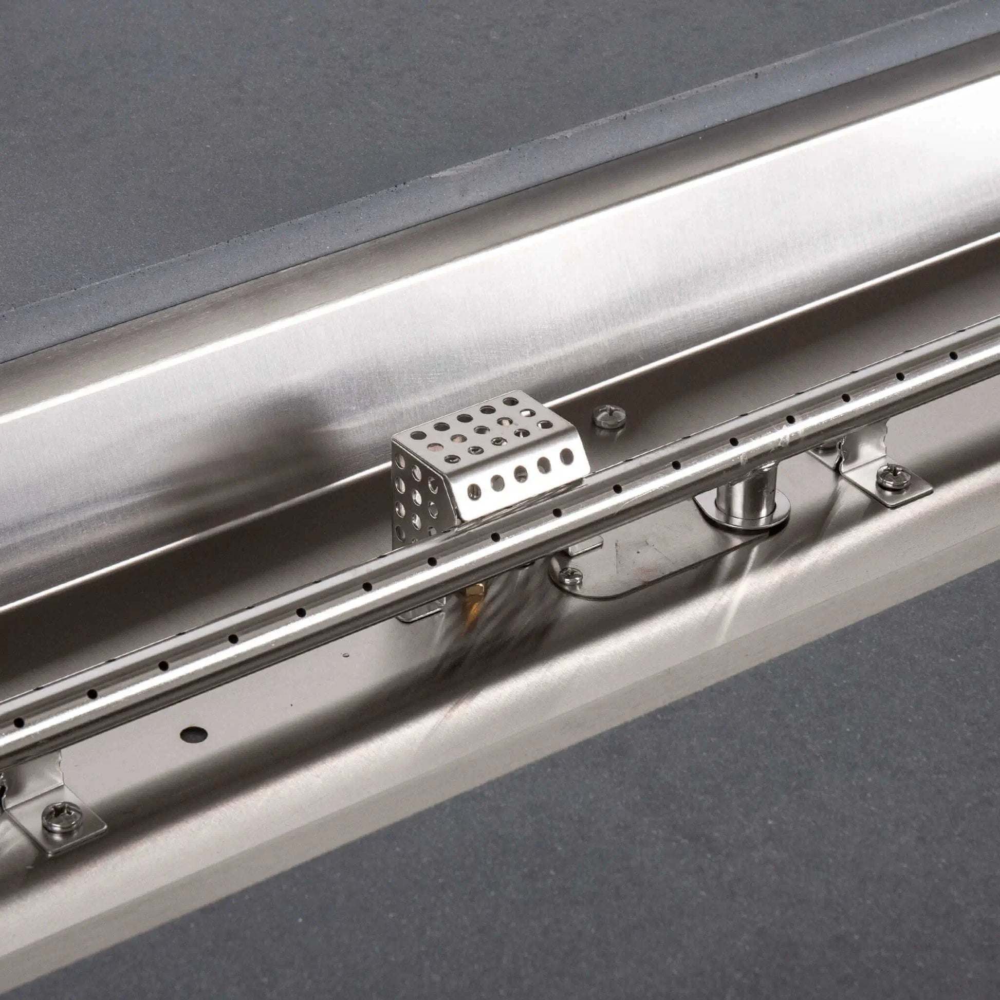 Detailed view inside the Flame Havens Refuge Series rectangular gray propane fire pit, showing the stainless steel linear burner tube with evenly spaced ports, reflective burner pan, support brackets, fasteners, and perforated CSA ignition cage.