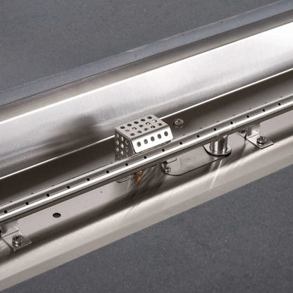 Detailed view inside the Flame Havens Refuge Series rectangular gray propane fire pit, showing the stainless steel linear burner tube with evenly spaced ports, reflective burner pan, support brackets, fasteners, and perforated CSA ignition cage.