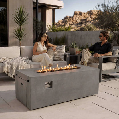 Couple relaxing with drinks around a lit Flame Havens Refuge Series 68 x 30 x 24-inch rectangular gray propane fire pit on a modern desert patio. Flames rise across tempered black fire glass, with the recessed controls and hidden tank access panel visible on the front and side.