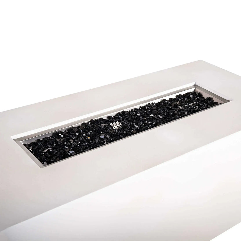 Angled close-up of the Flame Havens Refuge Series 68 x 30 x 24-inch rectangular propane gas fire pit in white GFRC concrete, showing the long stainless steel burner pan filled with reflective black fire glass and the protective ignition cage centered among the glass.