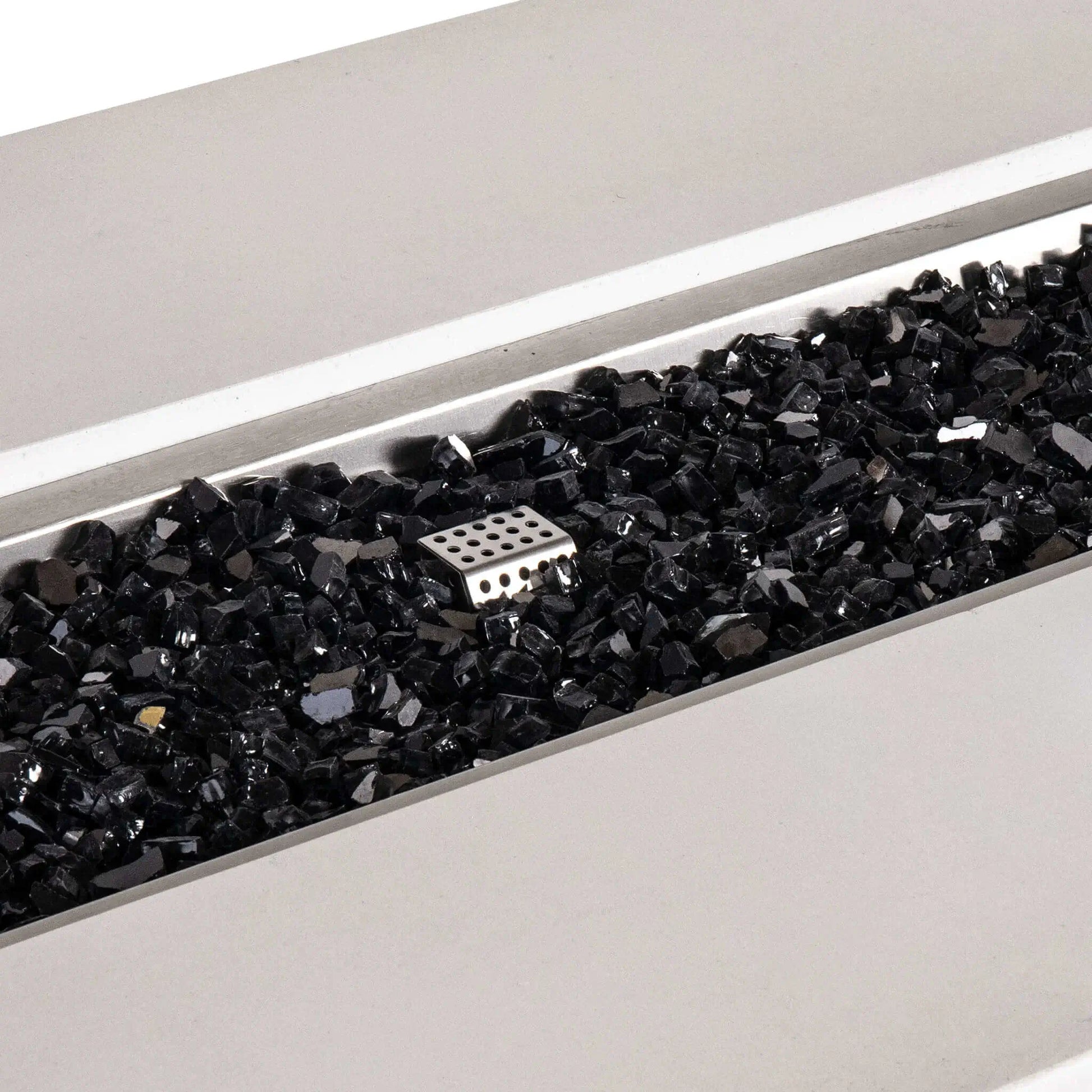 Close-up of the Flame Havens Refuge Series white GFRC concrete propane fire pit, highlighting the reflective black fire glass inside the stainless steel burner pan. A perforated metal ignition cage is visible among the glass in this detailed view of the 68 x 30 x 24-inch rectangular fire pit.