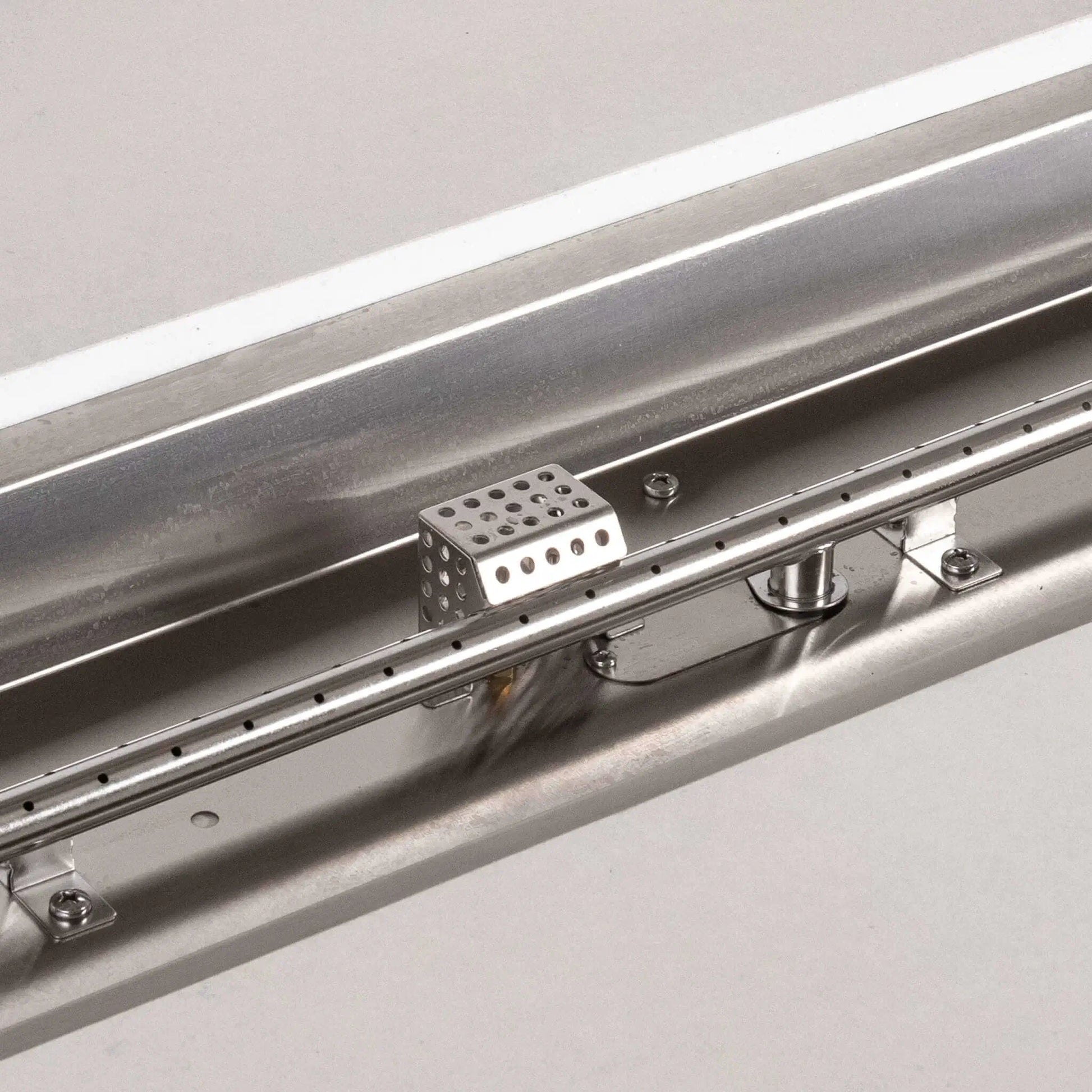 Detailed close-up of the Flame Havens Refuge Series stainless steel linear burner and CSA ignition components. The image highlights the evenly spaced burner ports, mounting hardware, central gas connection, reflective stainless steel pan, and protective perforated ignition cage inside the white propane fire pit.