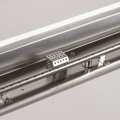 Detailed close-up of the Flame Havens Refuge Series stainless steel linear burner and CSA ignition components. The image highlights the evenly spaced burner ports, mounting hardware, central gas connection, reflective stainless steel pan, and protective perforated ignition cage inside the white propane fire pit.