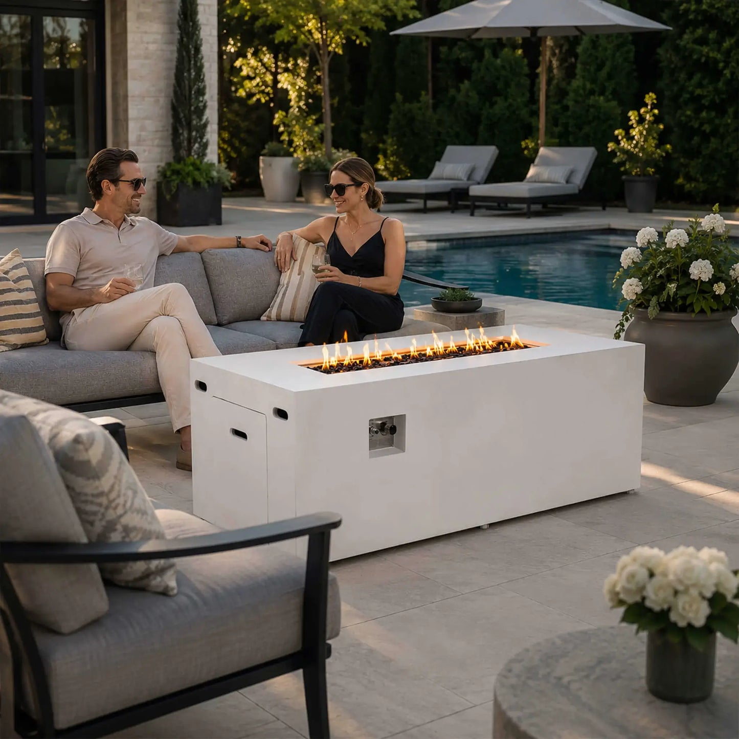 Couple relaxing beside a lit Flame Havens Refuge Series 68 x 30 x 24-inch white rectangular propane fire pit on an upscale poolside patio. Flames glow above tempered black fire glass, while the hidden tank door and recessed controls remain visible among modern seating, lounge chairs, flowers, and landscaped greenery.