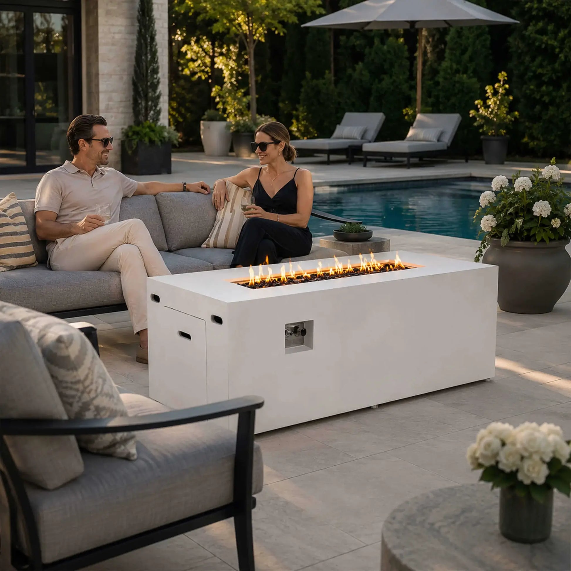 Couple relaxing beside a lit Flame Havens Refuge Series 68 x 30 x 24-inch white rectangular propane fire pit on an upscale poolside patio. Flames glow above tempered black fire glass, while the hidden tank door and recessed controls remain visible among modern seating, lounge chairs, flowers, and landscaped greenery.