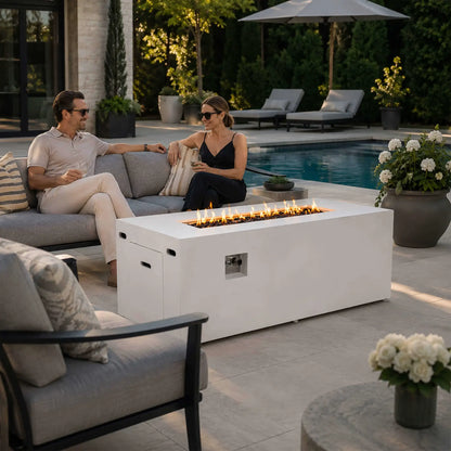 Couple relaxing beside a lit Flame Havens Refuge Series 68 x 30 x 24-inch white rectangular propane fire pit on an upscale poolside patio. Flames glow above tempered black fire glass, while the hidden tank door and recessed controls remain visible among modern seating, lounge chairs, flowers, and landscaped greenery.