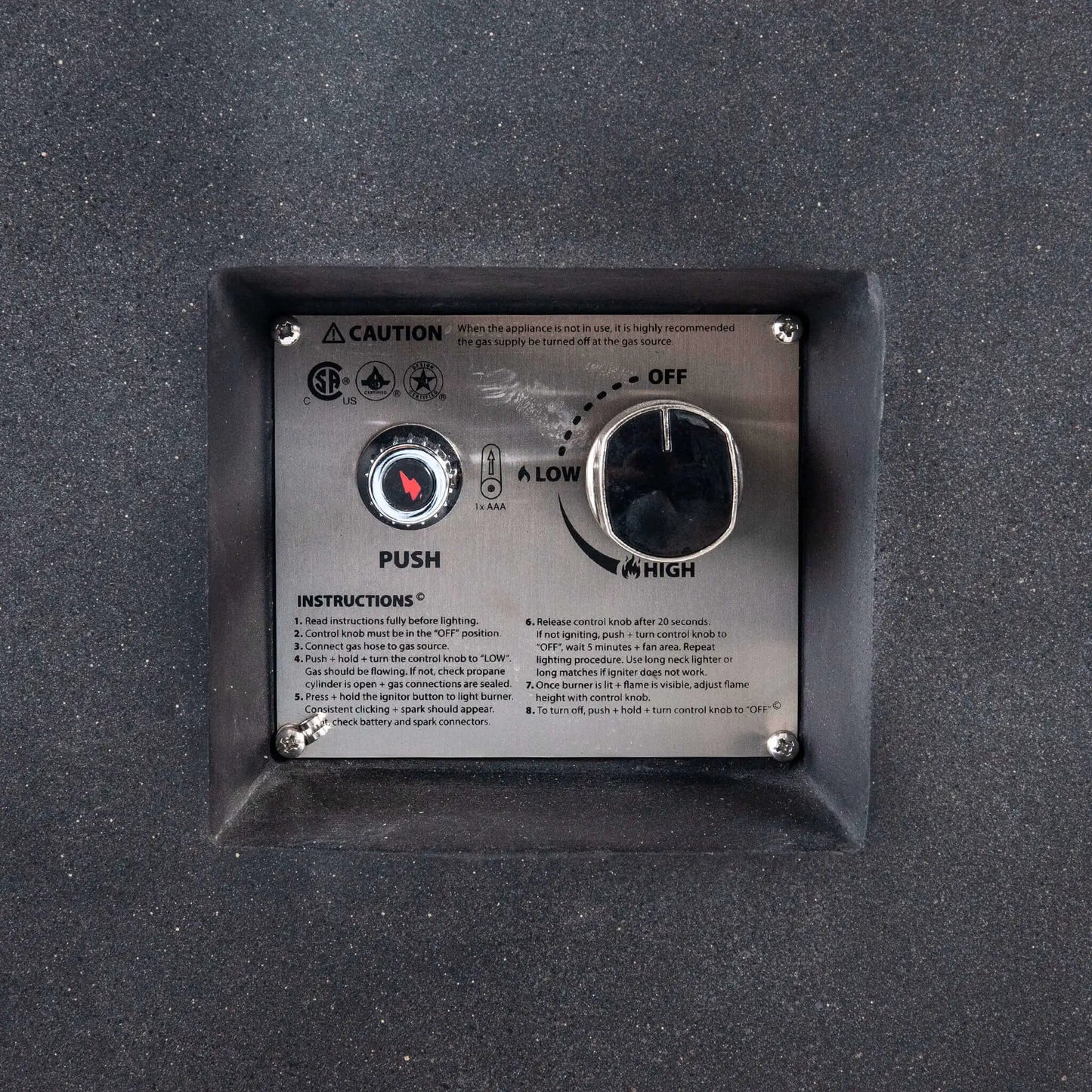 Close-up of the recessed CSA ignition controls on the Flame Havens Refuge Series 68 x 30 x 24-inch black GFRC concrete propane fire pit. The stainless steel panel shows a push-button spark ignitor, AAA battery indicator, adjustable control knob with off, low, and high settings, safety certifications, and operating instructions.