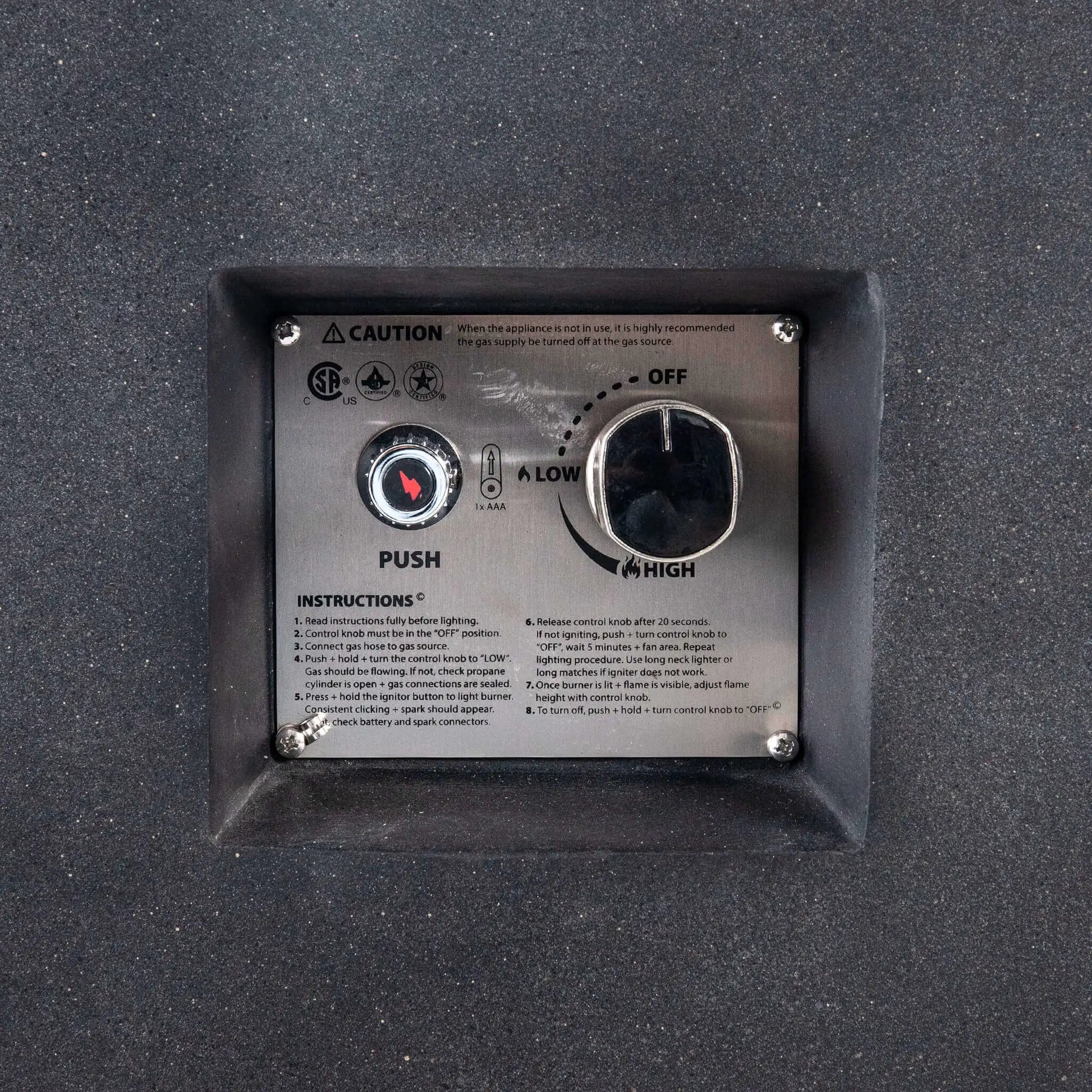 Close-up of the recessed CSA ignition controls on the Flame Havens Refuge Series 68 x 30 x 24-inch black GFRC concrete propane fire pit. The stainless steel panel shows a push-button spark ignitor, AAA battery indicator, adjustable control knob with off, low, and high settings, safety certifications, and operating instructions.