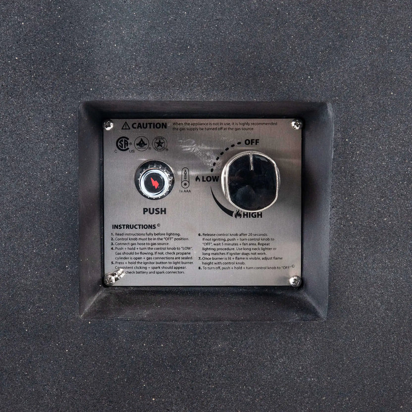 Close-up of the recessed CSA ignition controls on the Flame Havens Refuge Series 68 x 30 x 24-inch black GFRC concrete propane fire pit. The stainless steel panel shows a push-button spark ignitor, AAA battery indicator, adjustable control knob with off, low, and high settings, safety certifications, and operating instructions.
