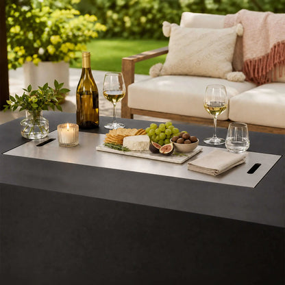 Flame Havens Refuge Series 68 x 30 x 24-inch black rectangular GFRC concrete propane fire pit with its stainless steel burner cover used as a tabletop. The outdoor lifestyle setting features wine, glasses, cheese, crackers, grapes, figs, a candle, greenery, and cushioned patio seating.