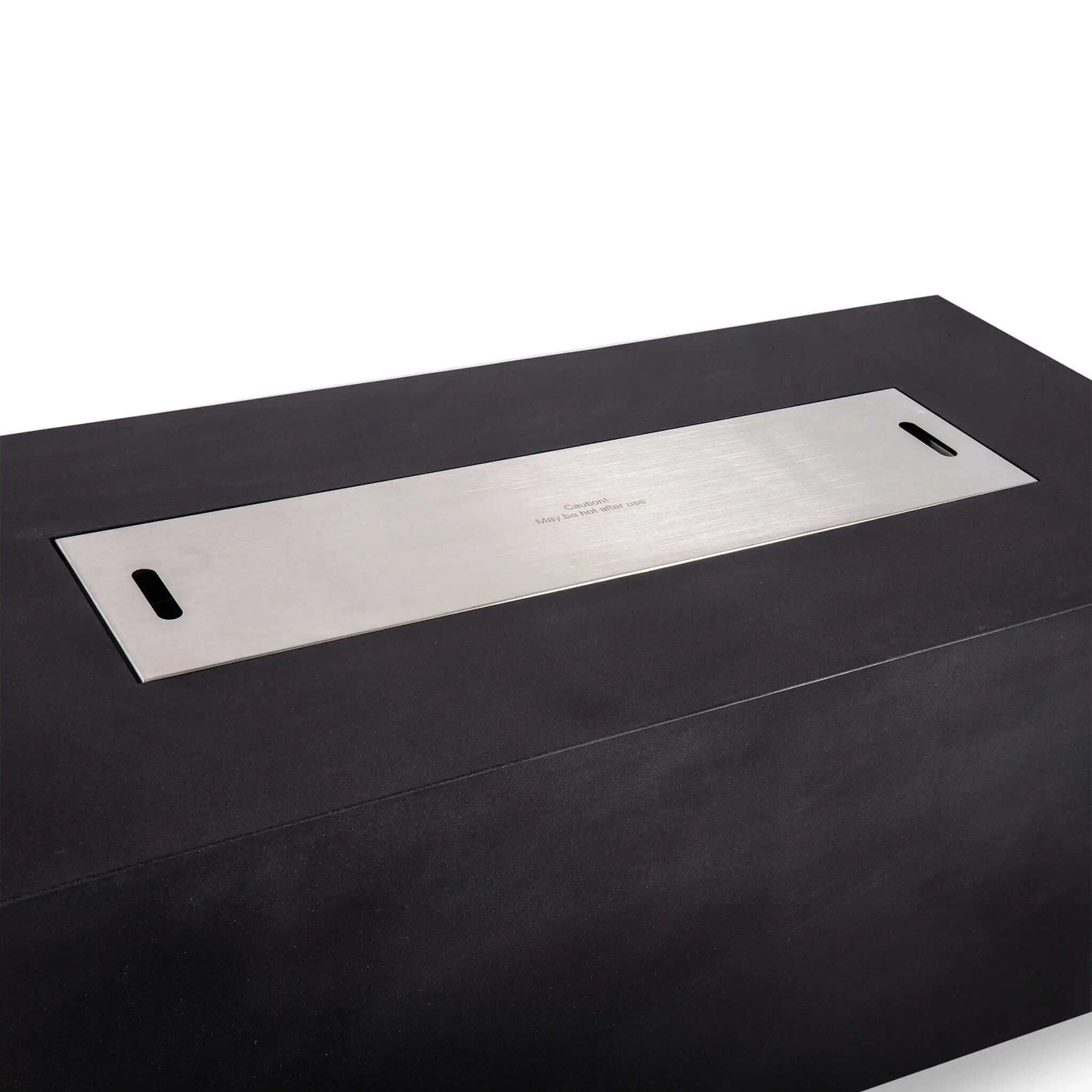 Stainless steel burner cover installed flush over the opening of the black Flame Havens Refuge Series 68 x 30 x 24-inch rectangular GFRC concrete propane fire pit, featuring two integrated lift handles and a centered caution marking for safe handling after use.