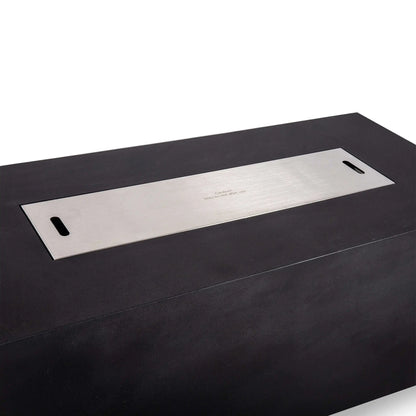 Stainless steel burner cover installed flush over the opening of the black Flame Havens Refuge Series 68 x 30 x 24-inch rectangular GFRC concrete propane fire pit, featuring two integrated lift handles and a centered caution marking for safe handling after use.