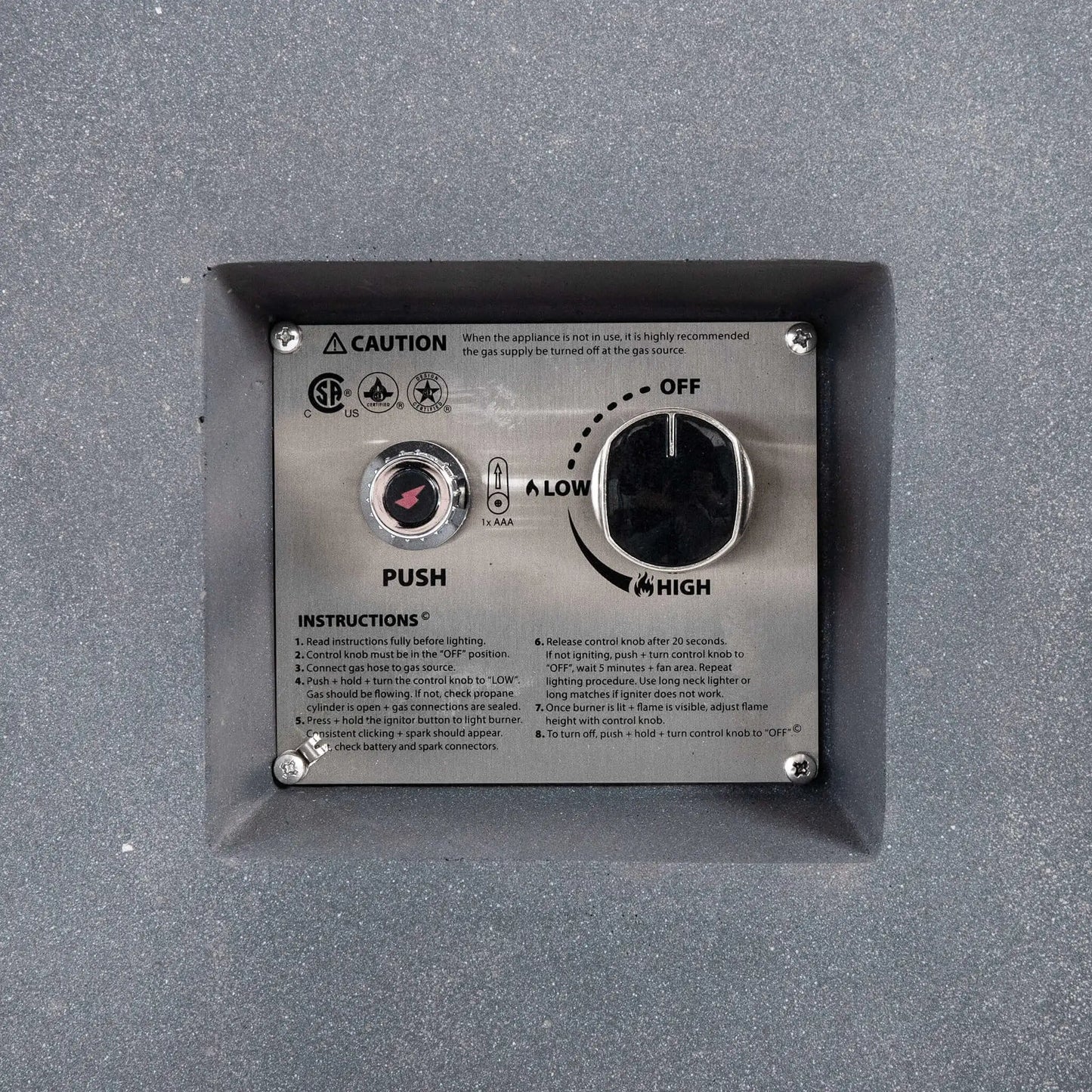 Close-up of the recessed CSA ignition controls on the Flame Havens Refuge Series gray propane gas fire pit, showing the push-button spark igniter, adjustable control knob with OFF, LOW, and HIGH settings, operating instructions, and safety markings on a stainless steel panel.