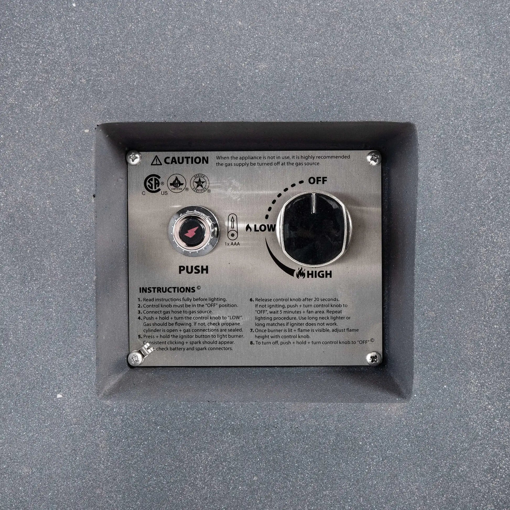 Close-up of the recessed CSA ignition controls on the Flame Havens Refuge Series gray propane gas fire pit, showing the push-button spark igniter, adjustable control knob with OFF, LOW, and HIGH settings, operating instructions, and safety markings on a stainless steel panel.