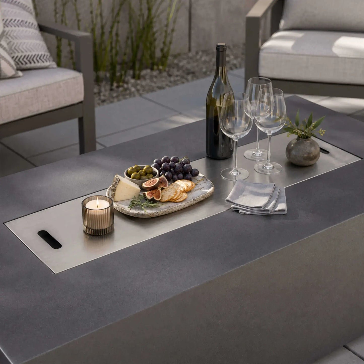 Stainless steel burner cover on the Flame Havens Refuge Series 68 x 30 x 24-inch gray propane fire pit used as a tabletop on a modern patio, arranged with wine, glasses, cheese, crackers, figs, grapes, olives, a candle, and a small vase.