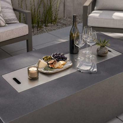 Stainless steel burner cover on the Flame Havens Refuge Series 68 x 30 x 24-inch gray propane fire pit used as a tabletop on a modern patio, arranged with wine, glasses, cheese, crackers, figs, grapes, olives, a candle, and a small vase.