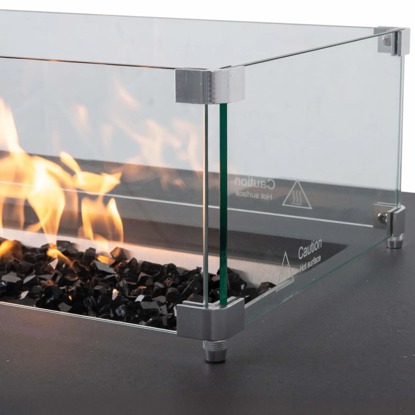 Close-up of the tempered glass wind guard on a Flame Havens Refuge Series 68 x 30 x 24-inch gray propane fire pit, with flames burning over reflective black fire glass. Stainless steel corner brackets and raised feet secure the clear glass panels around the burner.