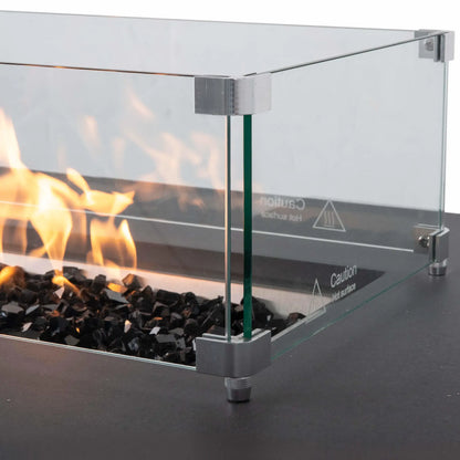 Close-up of the tempered glass wind guard on a Flame Havens Refuge Series 68 x 30 x 24-inch gray propane fire pit, with flames burning over reflective black fire glass. Stainless steel corner brackets and raised feet secure the clear glass panels around the burner.