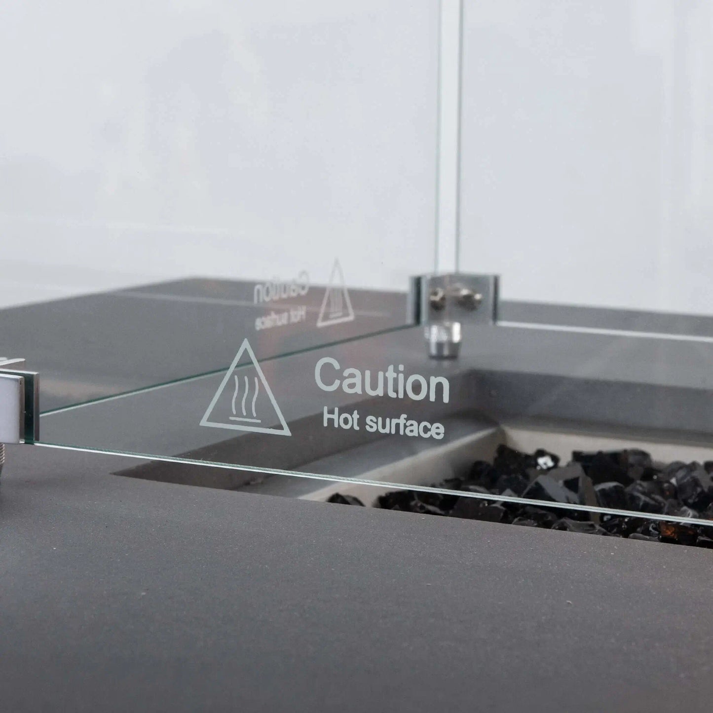 Detailed close-up of the Flame Havens Refuge Series tempered glass wind guard, focusing on the etched “Caution Hot Surface” warning, clear glass panel, stainless steel corner hardware, and black fire glass inside the gray rectangular propane fire pit.