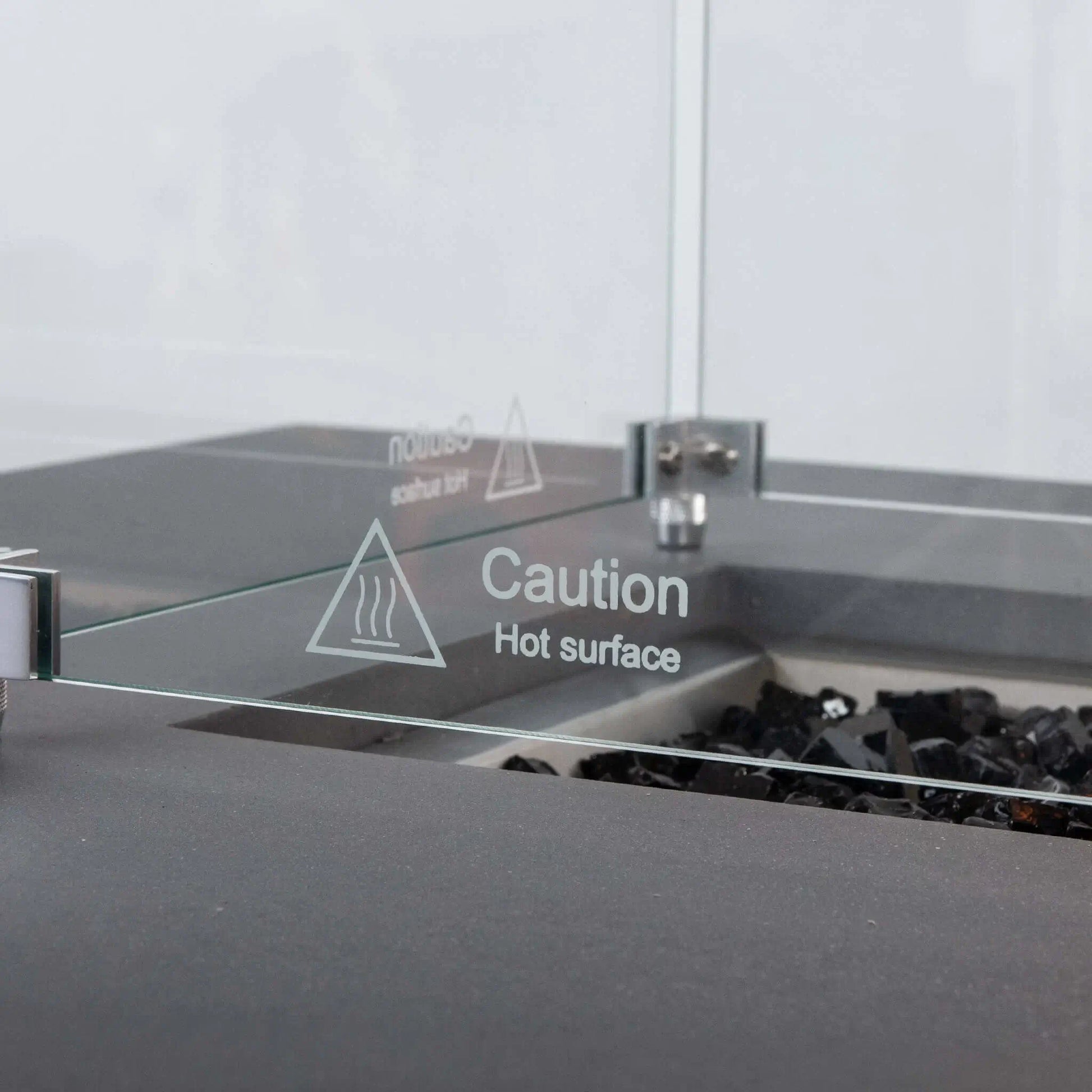 Detailed close-up of the Flame Havens Refuge Series tempered glass wind guard, focusing on the etched “Caution Hot Surface” warning, clear glass panel, stainless steel corner hardware, and black fire glass inside the gray rectangular propane fire pit.