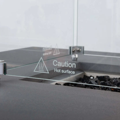 Detailed close-up of the Flame Havens Refuge Series tempered glass wind guard, focusing on the etched “Caution Hot Surface” warning, clear glass panel, stainless steel corner hardware, and black fire glass inside the gray rectangular propane fire pit.