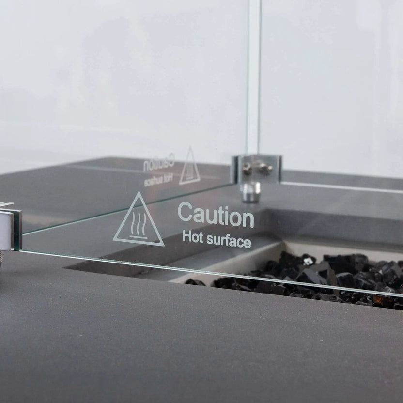 Detailed close-up of the Flame Havens Refuge Series tempered glass wind guard, focusing on the etched “Caution Hot Surface” warning, clear glass panel, stainless steel corner hardware, and black fire glass inside the gray rectangular propane fire pit.