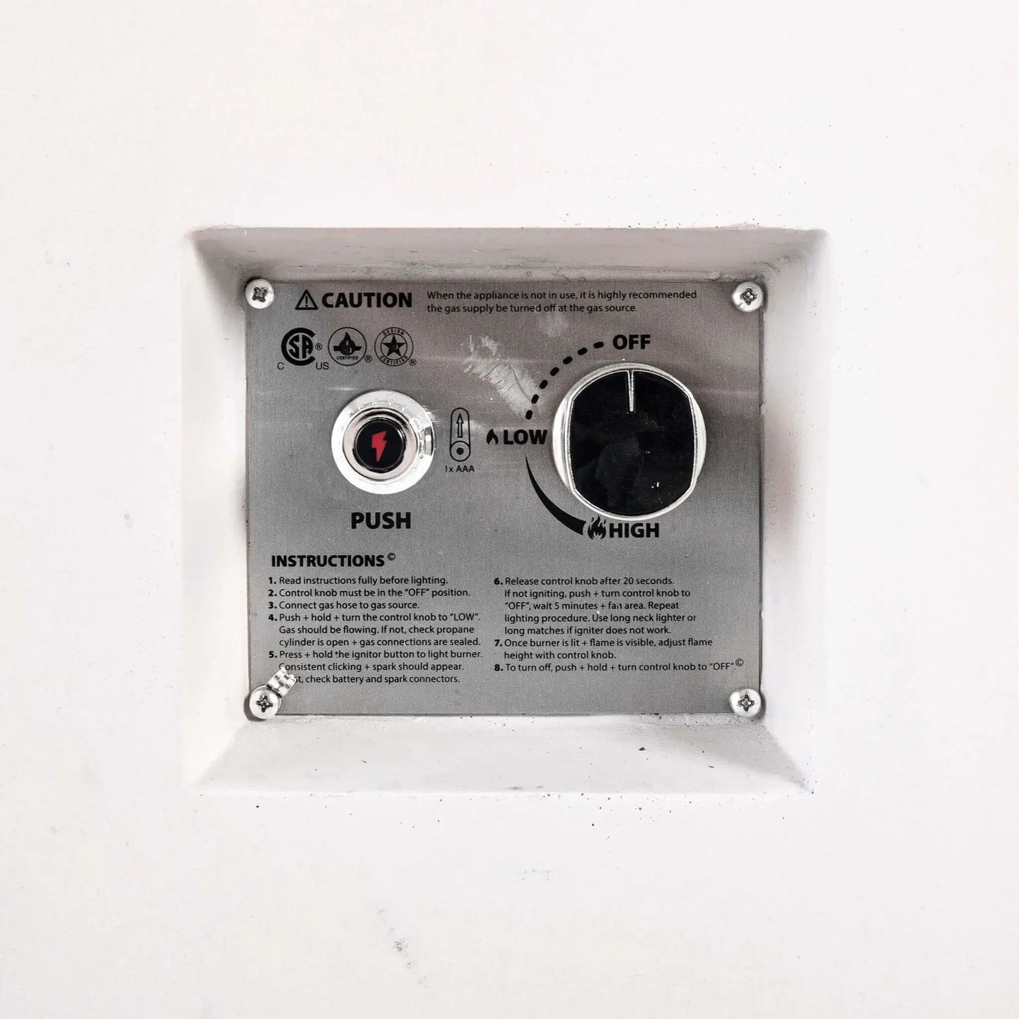 Close-up of the recessed CSA ignition controls on the Flame Havens Refuge Series 68 x 30 x 24-inch white rectangular propane fire pit. The stainless steel panel includes a push-button battery ignitor, large flame control knob with off, low, and high settings, safety certifications, caution notice, and lighting instructions.