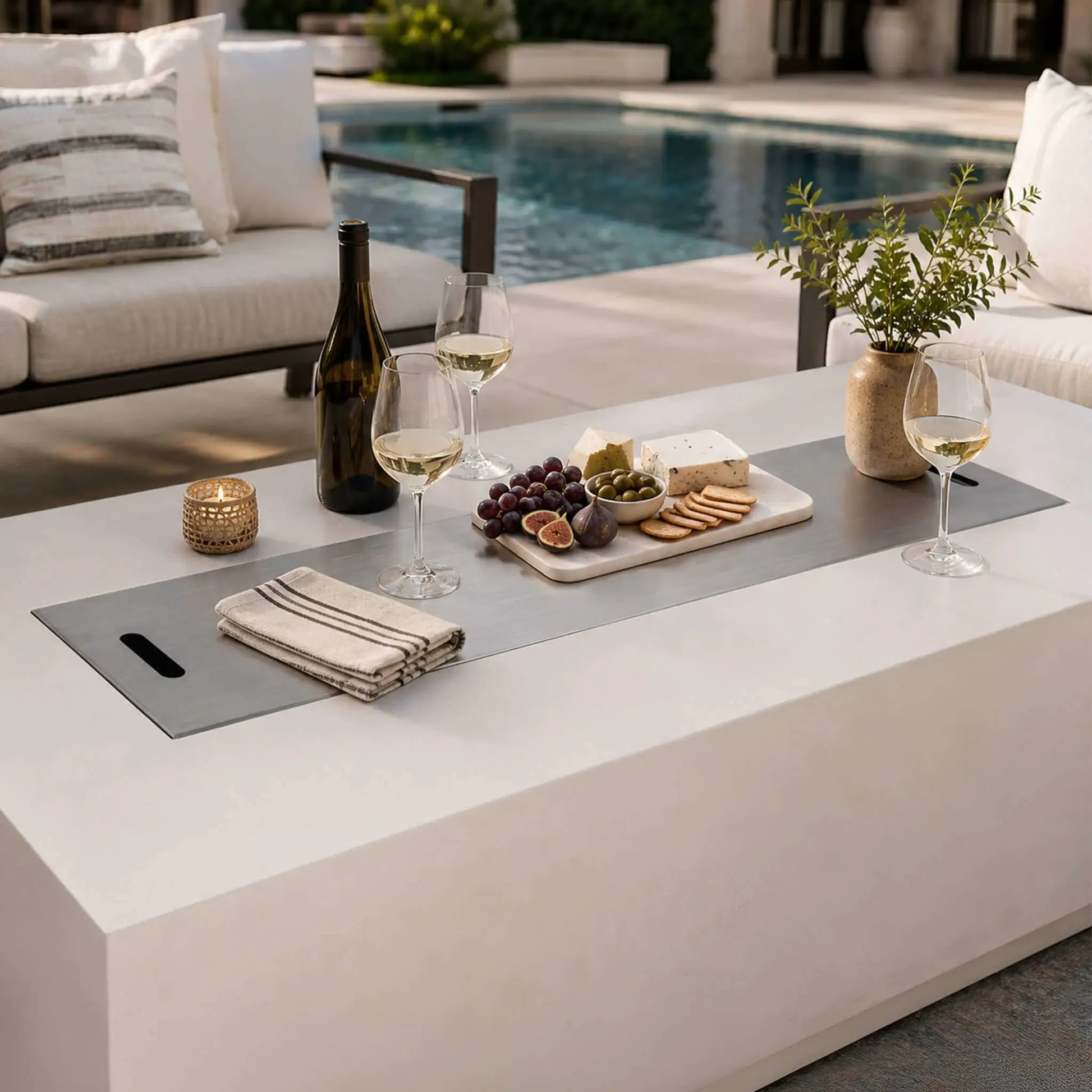 Flame Havens Refuge Series 68 x 30 x 24-inch white GFRC propane fire pit with its stainless steel burner cover used as a poolside tabletop. Wine glasses, a bottle, candle, napkins, flowers, and a charcuterie board with cheese, grapes, figs, olives, and crackers create an inviting outdoor entertaining scene.
