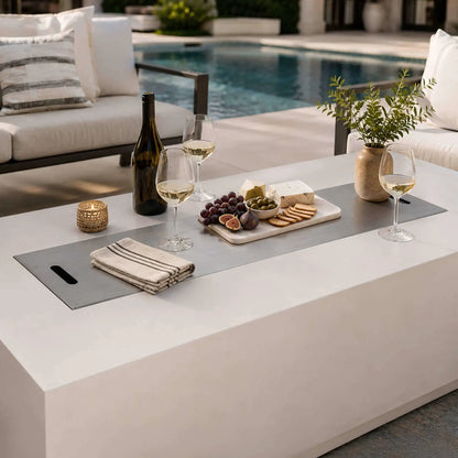 Flame Havens Refuge Series 68 x 30 x 24-inch white GFRC propane fire pit with its stainless steel burner cover used as a poolside tabletop. Wine glasses, a bottle, candle, napkins, flowers, and a charcuterie board with cheese, grapes, figs, olives, and crackers create an inviting outdoor entertaining scene.