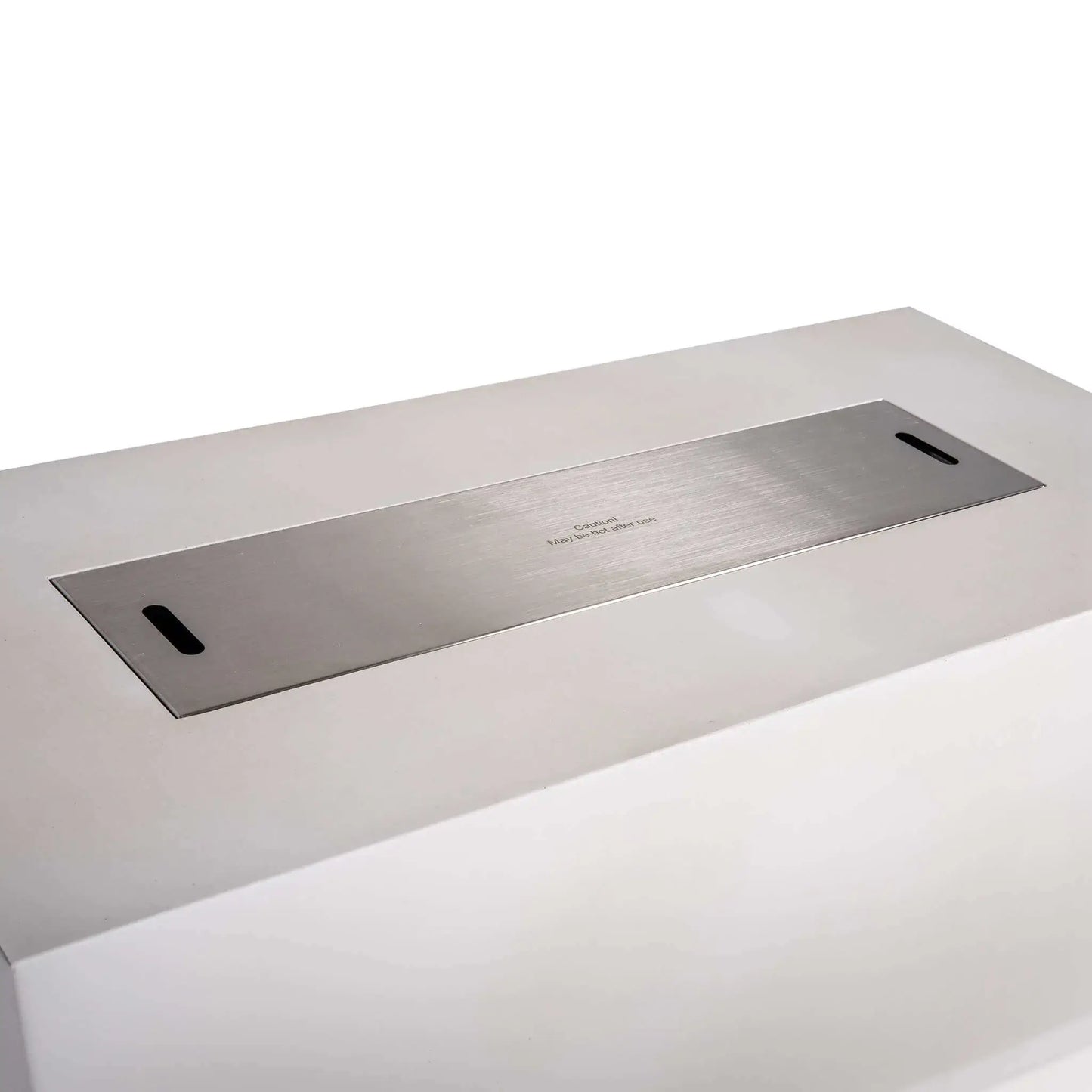 Close angled view of a stainless steel burner cover fitted over the opening of the Flame Havens Refuge Series 68 x 30 x 24-inch white rectangular propane fire pit. The brushed metal cover sits nearly flush with the GFRC concrete top and includes two hand slots for easy removal and a centered hot-surface caution notice.