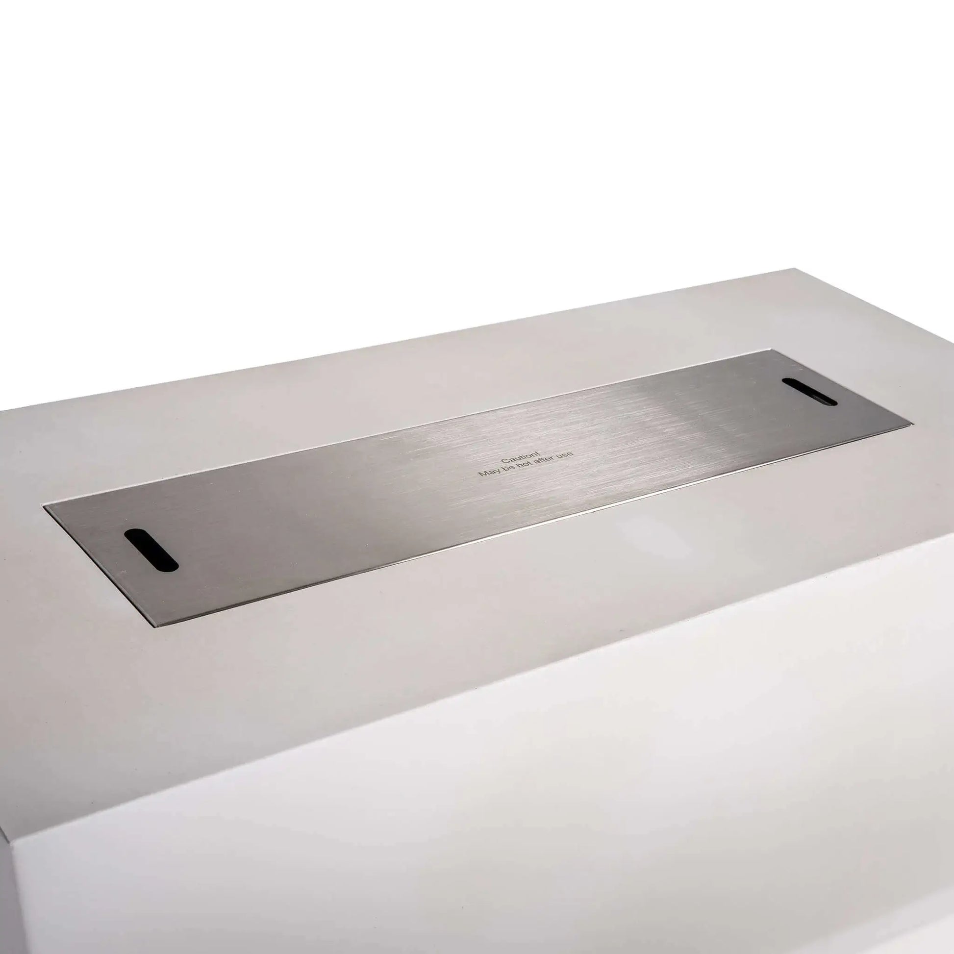 Close angled view of a stainless steel burner cover fitted over the opening of the Flame Havens Refuge Series 68 x 30 x 24-inch white rectangular propane fire pit. The brushed metal cover sits nearly flush with the GFRC concrete top and includes two hand slots for easy removal and a centered hot-surface caution notice.