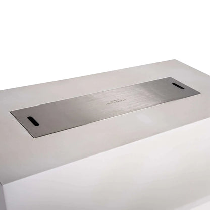 Close angled view of a stainless steel burner cover fitted over the opening of the Flame Havens Refuge Series 68 x 30 x 24-inch white rectangular propane fire pit. The brushed metal cover sits nearly flush with the GFRC concrete top and includes two hand slots for easy removal and a centered hot-surface caution notice.