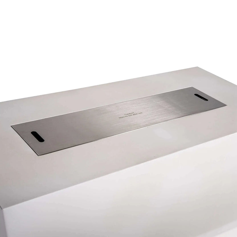 Close angled view of a stainless steel burner cover fitted over the opening of the Flame Havens Refuge Series 68 x 30 x 24-inch white rectangular propane fire pit. The brushed metal cover sits nearly flush with the GFRC concrete top and includes two hand slots for easy removal and a centered hot-surface caution notice.
