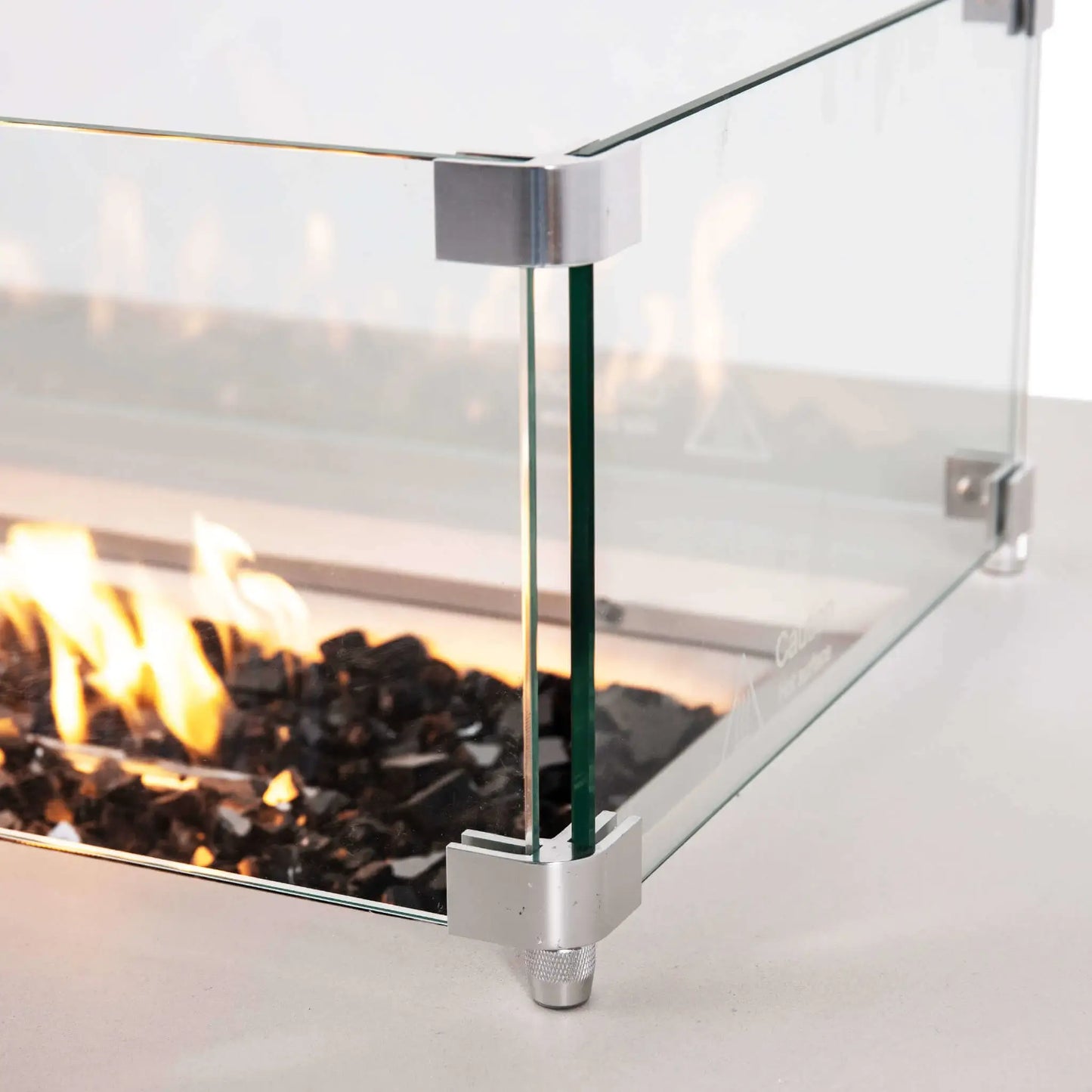 Close-up of the tempered glass wind guard surrounding the lit burner on a Flame Havens Refuge Series white rectangular propane fire pit. Clear glass panels are joined with brushed metal corner brackets and raised feet, while bright flames glow over tempered black fire glass inside the 68 x 30 x 24-inch fire pit.