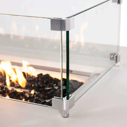 Close-up of the tempered glass wind guard surrounding the lit burner on a Flame Havens Refuge Series white rectangular propane fire pit. Clear glass panels are joined with brushed metal corner brackets and raised feet, while bright flames glow over tempered black fire glass inside the 68 x 30 x 24-inch fire pit.