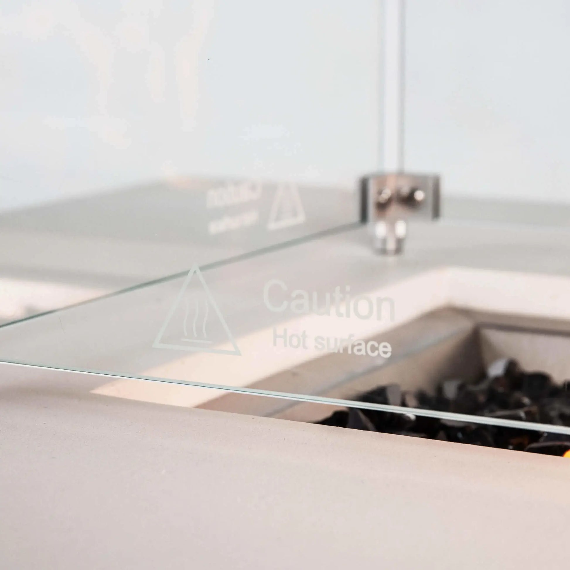 Detailed view of the tempered glass wind guard on the Flame Havens Refuge Series 68 x 30 x 24-inch white propane fire pit. The clear panel features an etched hot-surface caution symbol, with stainless steel corner hardware and tempered black fire glass visible behind it inside the recessed burner area.