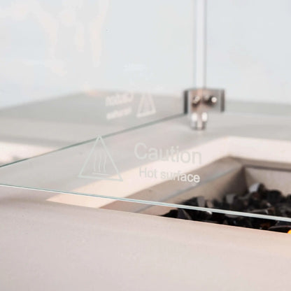 Detailed view of the tempered glass wind guard on the Flame Havens Refuge Series 68 x 30 x 24-inch white propane fire pit. The clear panel features an etched hot-surface caution symbol, with stainless steel corner hardware and tempered black fire glass visible behind it inside the recessed burner area.