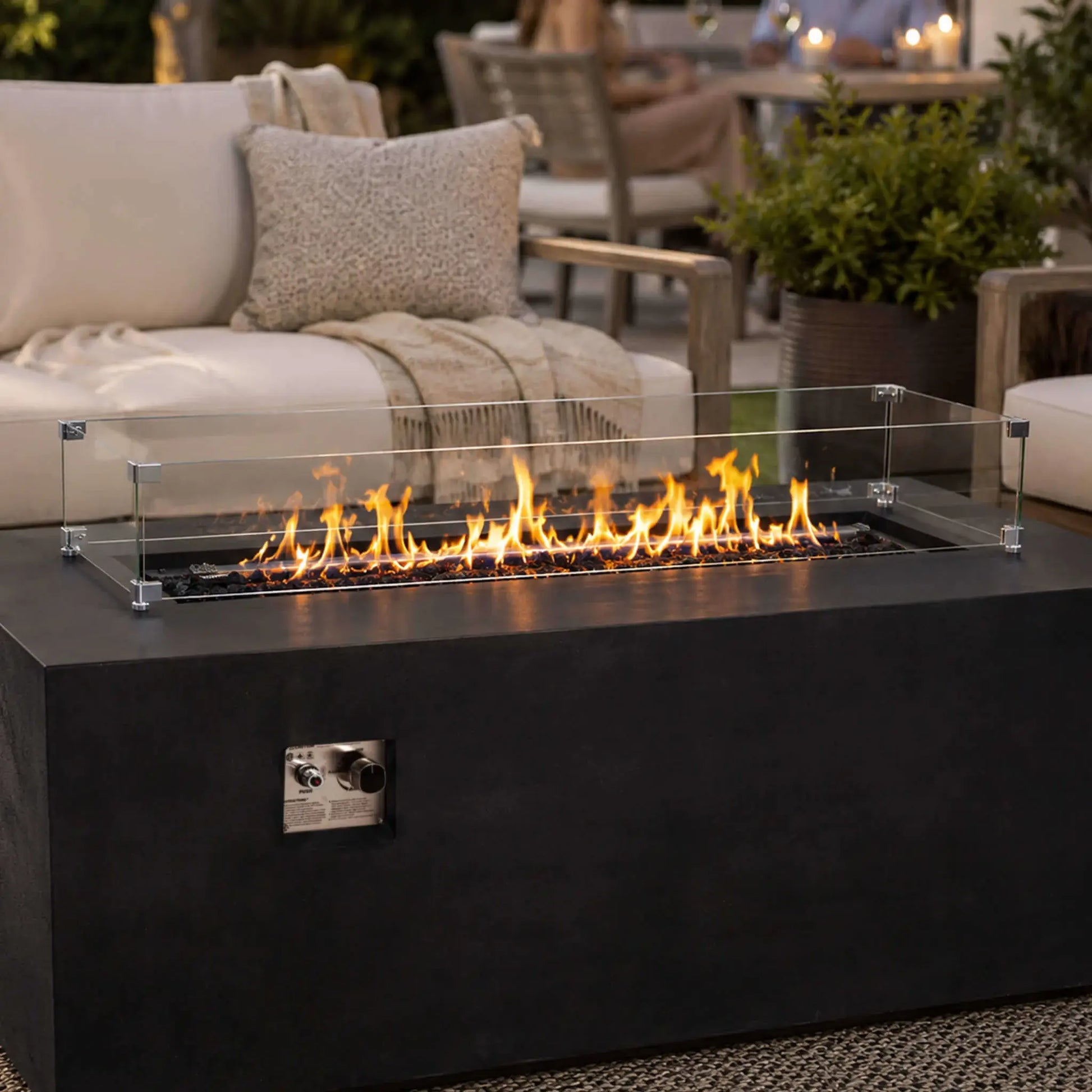 Flame Havens Refuge Series 68 x 30 x 24-inch rectangular black GFRC concrete propane fire pit burning in an inviting outdoor lounge. Tempered black fire glass and a clear glass wind guard frame the linear flames, with cushioned patio seating, pillows, a throw blanket, greenery, and guests in the softly lit background.