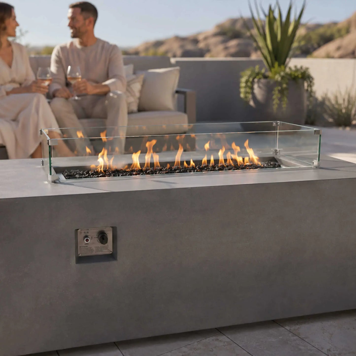 Flame Havens Refuge Series 68 x 30 x 24-inch rectangular gray propane fire pit lit on a modern outdoor patio, with flames rising through tempered black fire glass inside a clear wind guard while a couple relaxes with drinks in the background.