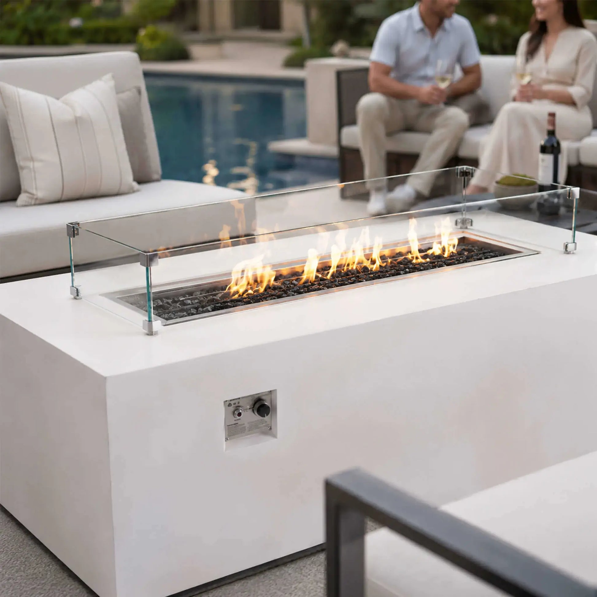 Flame Havens Refuge Series 68 x 30 x 24-inch white rectangular propane fire pit burning beside a residential pool. Flames rise over tempered black fire glass within a clear glass wind guard, while a couple enjoys wine on cushioned patio seating in the softly focused background.
