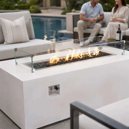 Flame Havens Refuge Series 68 x 30 x 24-inch white rectangular propane fire pit burning beside a residential pool. Flames rise over tempered black fire glass within a clear glass wind guard, while a couple enjoys wine on cushioned patio seating in the softly focused background.