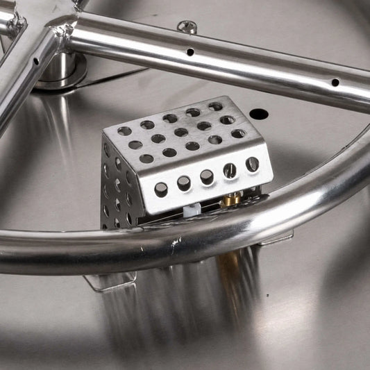 Close-up of a Flame Havens Refuge Series ignition shield installed inside a stainless steel fire pit burner pan. The perforated metal enclosure surrounds the igniter assembly beside the round burner tube, with portions of the electrode and brass fitting visible through the lower opening.