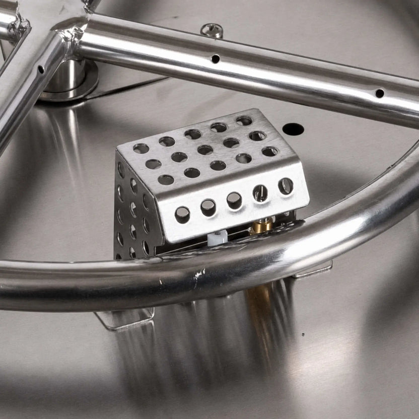 Close-up of a Flame Havens Refuge Series ignition shield installed inside a stainless steel fire pit burner pan. The perforated metal enclosure surrounds the igniter assembly beside the round burner tube, with portions of the electrode and brass fitting visible through the lower opening.