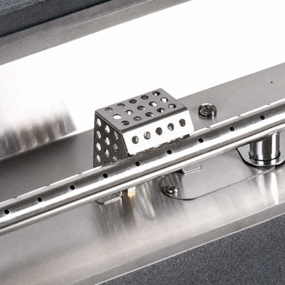 Flame Havens Refuge Series ignition shield installed in a stainless steel fire pit burner pan. The perforated protective cover sits directly behind the linear burner tube and its evenly spaced gas ports, showing how the shield is positioned around the ignition components within the burner assembly.