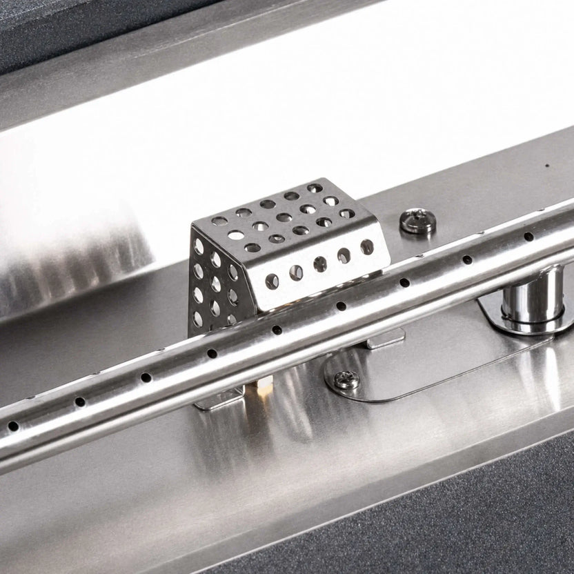 Flame Havens Refuge Series ignition shield installed in a stainless steel fire pit burner pan. The perforated protective cover sits directly behind the linear burner tube and its evenly spaced gas ports, showing how the shield is positioned around the ignition components within the burner assembly.