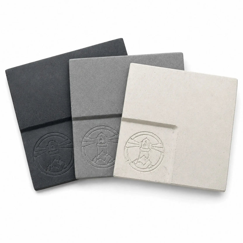 Three overlapping Flame Havens Refuge and Serenity Series GFRC concrete samples displaying the available Graphite Black, Natural Gray, and Ivory White finishes from left to right. Each square glass fiber reinforced concrete swatch has a lightly textured surface, rounded edges, and an embossed Flame Havens emblem near the lower corner.