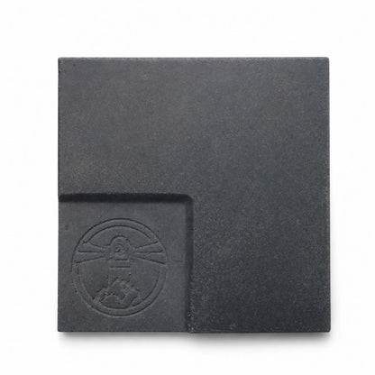 Flame Havens Refuge and Serenity Series GFRC glass fiber reinforced concrete sample in Graphite Black. The square finish swatch has a deep charcoal-black color, fine concrete texture, subtle tonal variation, softly rounded edges, and a recessed lower-left panel featuring an embossed Flame Havens emblem.
