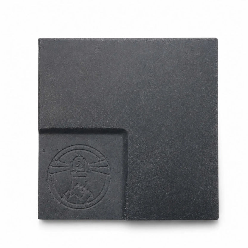 Flame Havens Refuge and Serenity Series GFRC glass fiber reinforced concrete sample in Graphite Black. The square finish swatch has a deep charcoal-black color, fine concrete texture, subtle tonal variation, softly rounded edges, and a recessed lower-left panel featuring an embossed Flame Havens emblem.